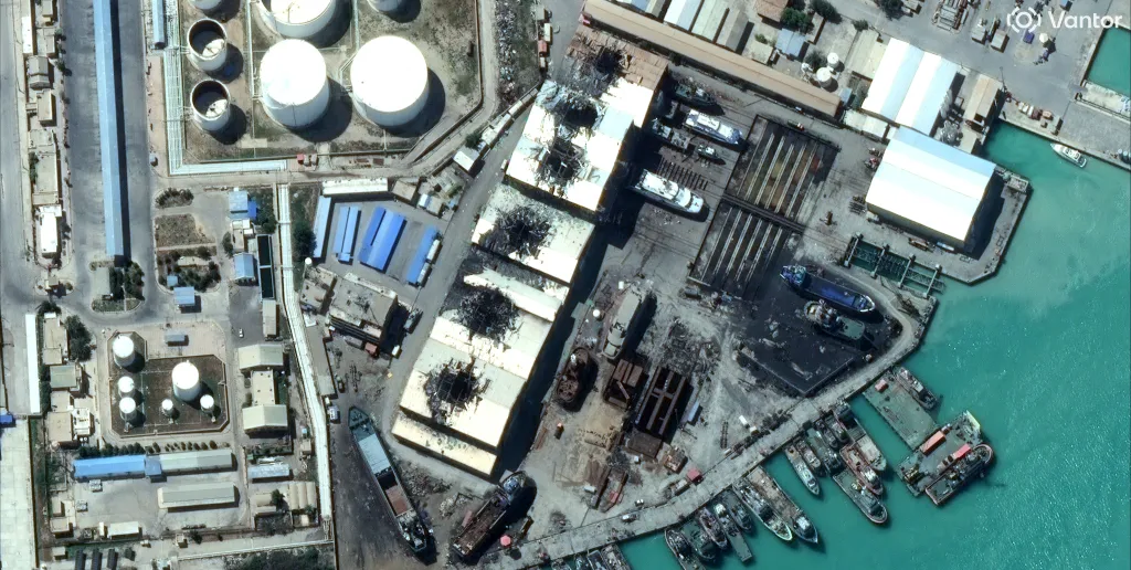 An Iranian naval base in Bushehr shows significant damage. A row of large warehouses runs across the center of the photo, and each warehouse has an obvious bomb entry hole in the center of the roof.